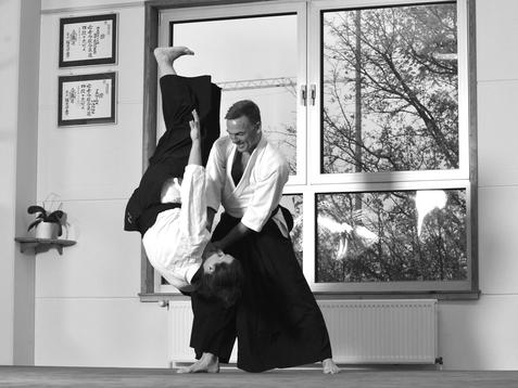 Zwei Kampfsportler führen eine Aikido-Technik in einem Dojo mit Fenster und Wänden voller Urkunden aus.