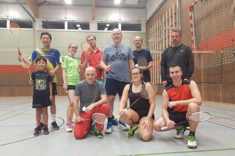 Eine Gruppe von 10 Badmintonspielern hat sich zu einem Gruppenfoto aufgestellt und hält dabei ihre Schläger hoch.