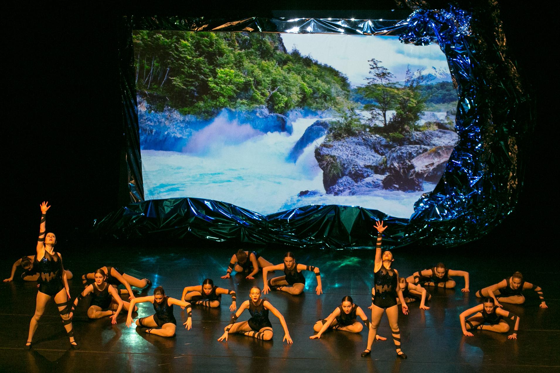 AQUA - Vom Regentropfen bis zum Ozean Balletttänzerinnen auf einer Bühne. Zwei stehen im Vordergrund und strecken einen Arm in die Höhe. Dahinter tanzen 14 Tänzerinnen am Boden. An die Rückwand ist ein Wasserfall.