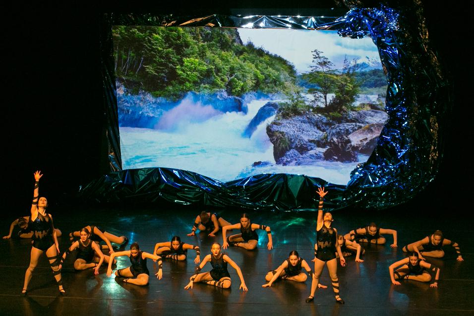 Balletttänzerinnen auf einer Bühne. Zwei stehen im Vordergrund und strecken einen Arm in die Höhe. Dahinter tanzen 14 Tänzerinnen am Boden. An die Rückwand ist ein Wasserfall.