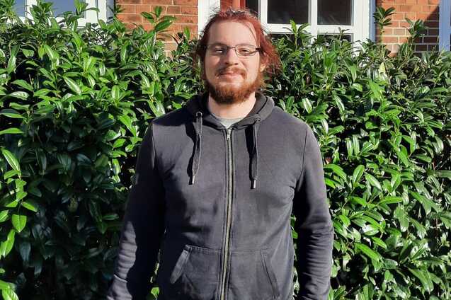 Porträt von Lukas Heinelt vor einer grünen Hecke.