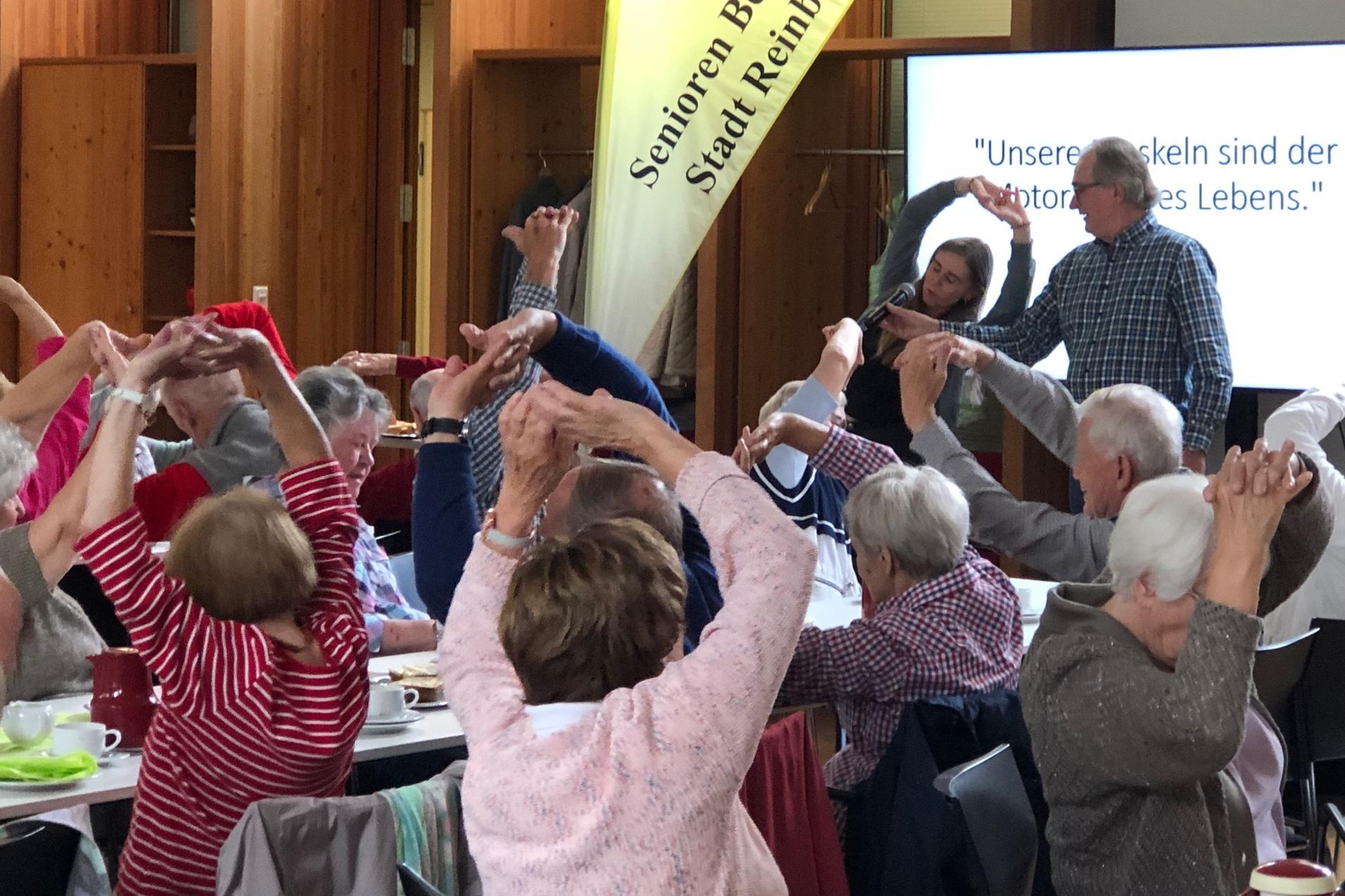 Vortrag Kraftsport im Alter Senioren sitzen an Tischen und strecken ihre Arme in die Luft zur Dehnung