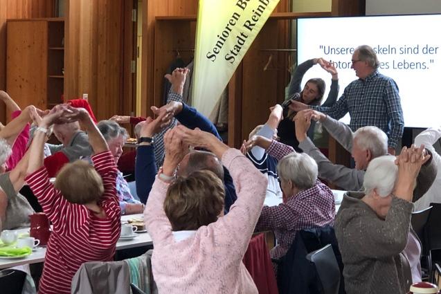 Senioren sitzen an Tischen und strecken ihre Arme in die Luft zur Dehnung