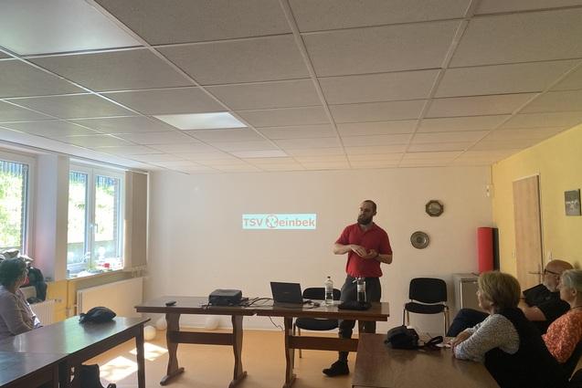 Personen sitzen um einen Tischkreis und schauen zum Referenten.