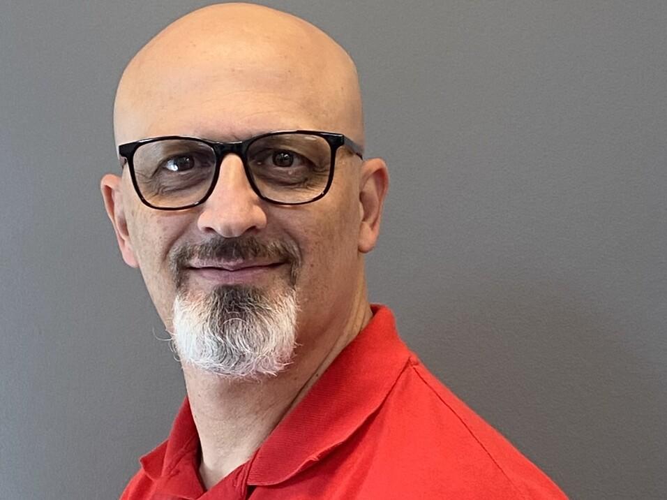 Mann mit Glatze und Brille trägt ein rotes Polohemd, lächelt vor einer grauen Wand. Namensschild sichtbar.