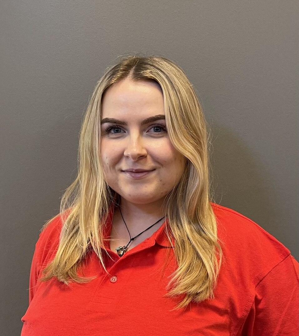 Porträt einer lächelnden Frau mit langen, blonden Haaren in einem roten Polohemd vor einem grauen Hintergrund.