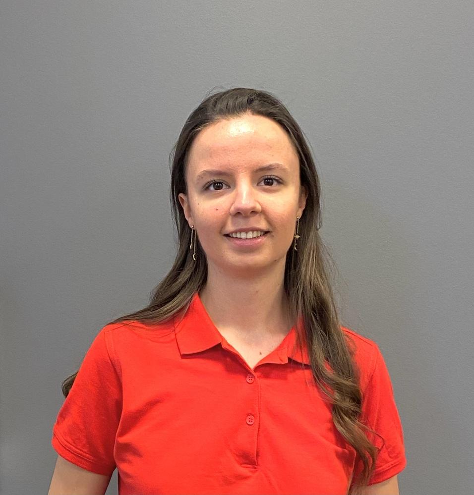 Junge Frau mit langen Haaren in rotem Shirt vor grauer Wand, lächelt in die Kamera.