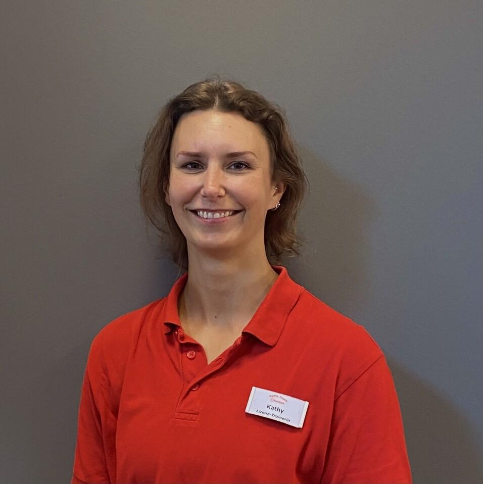 Porträt einer Frau mit kurzen Haaren in rotem Polo-Shirt, lächelnd vor einem grauen Hintergrund.