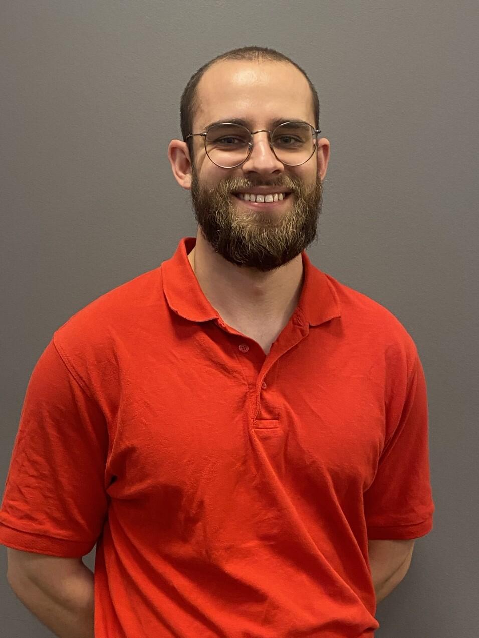 Porträt eines lächelnden Mannes mit Brille, in rotem Poloshirt vor grauem Hintergrund.