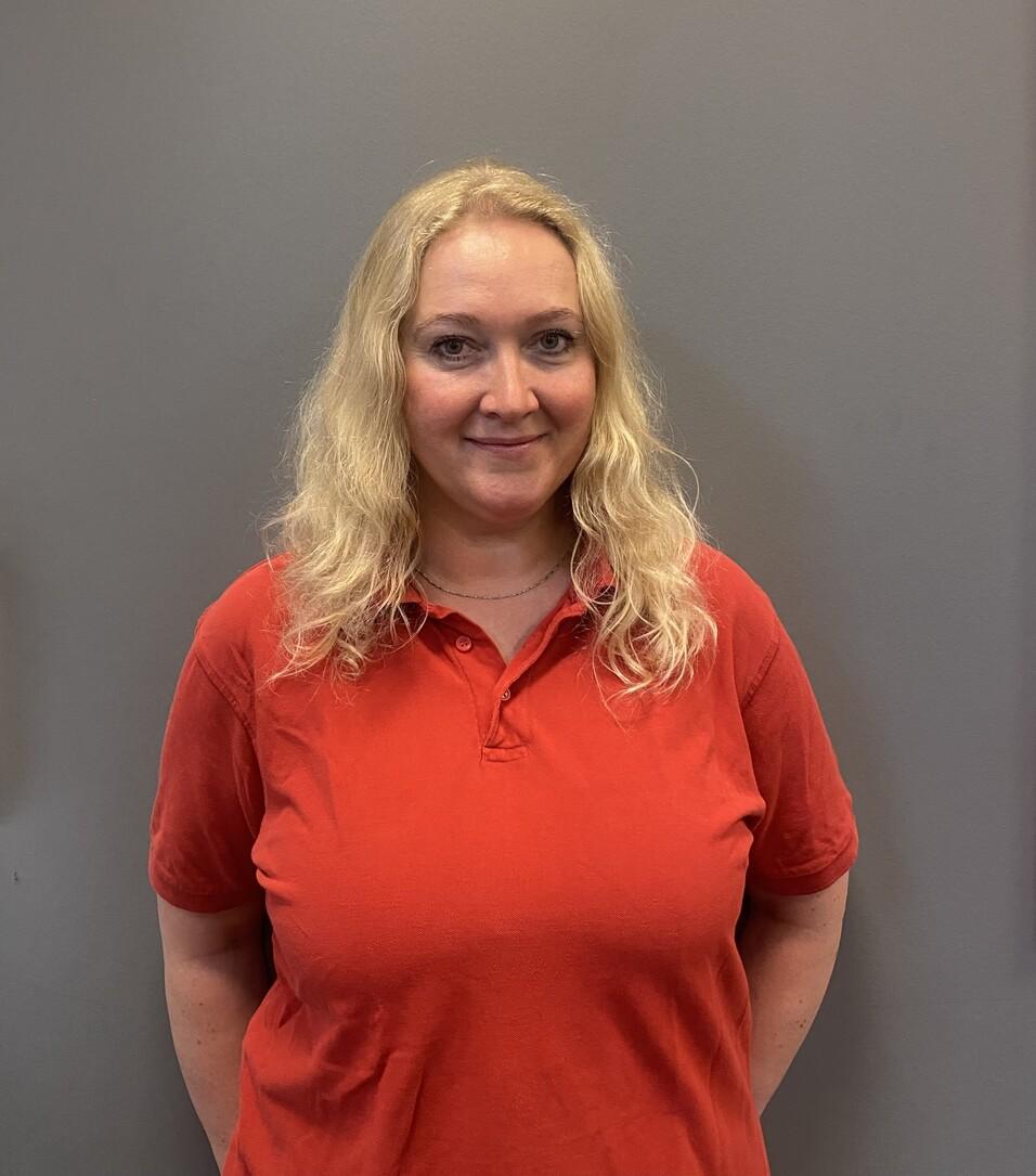 Frau mit langen blonden Haaren trägt ein rotes Polo-Shirt vor einem grauen Hintergrund.