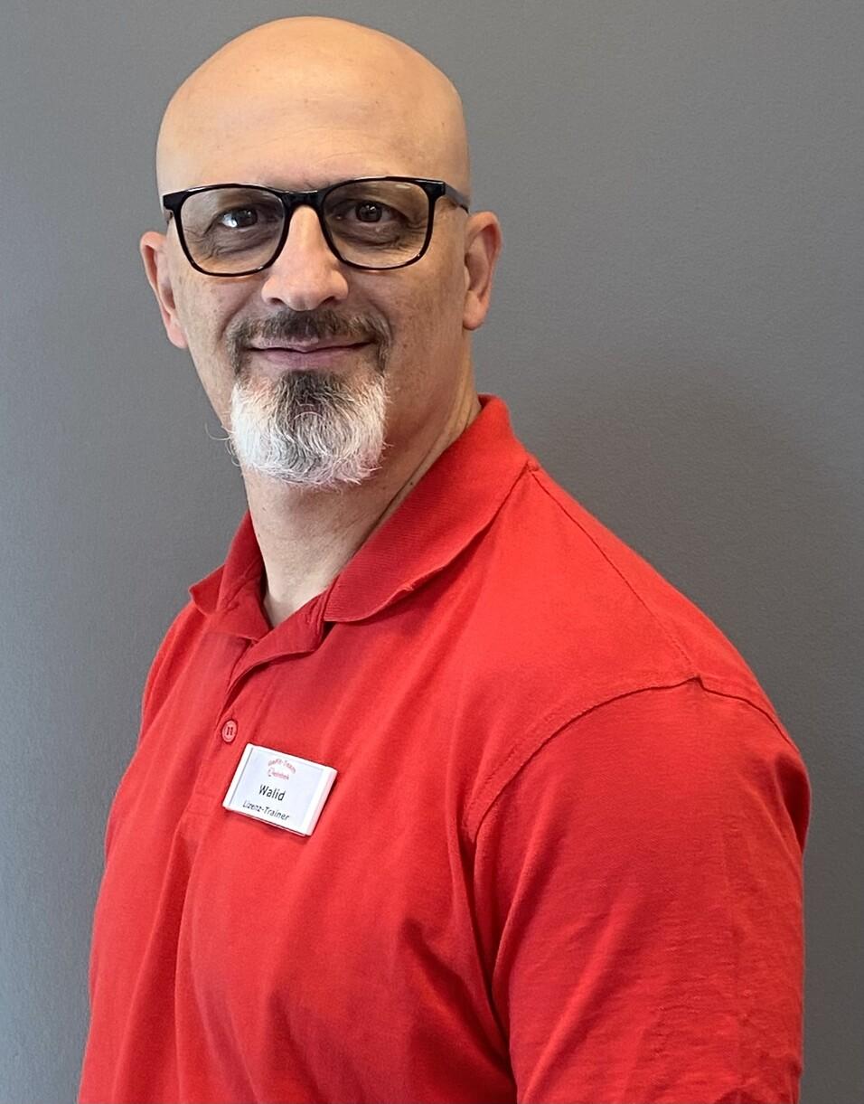 Mann mit Glatze und Brille trägt ein rotes Polohemd, lächelt vor einer grauen Wand. Namensschild sichtbar.