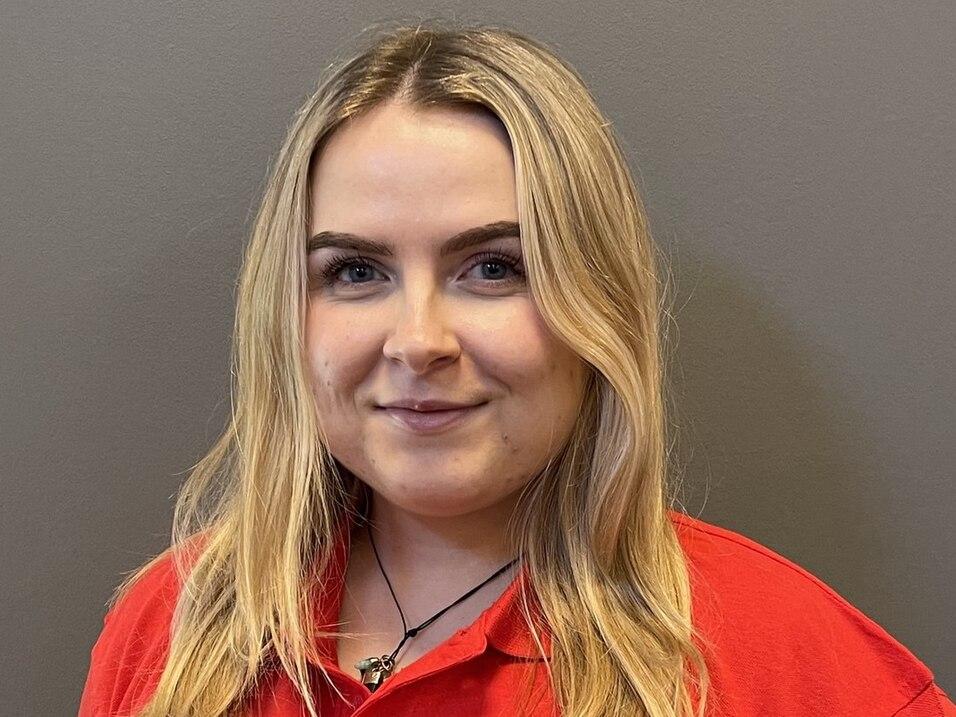 Porträt einer lächelnden Frau mit langen, blonden Haaren in einem roten Polohemd vor einem grauen Hintergrund.
