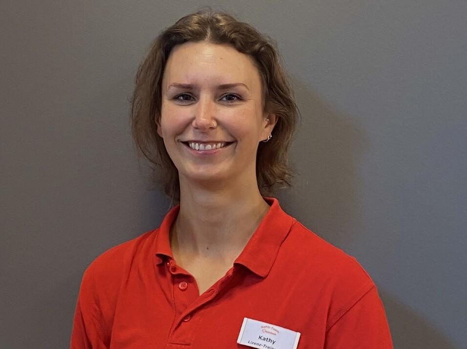 Porträt einer Frau mit kurzen Haaren in rotem Polo-Shirt, lächelnd vor einem grauen Hintergrund.