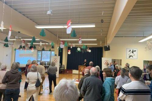 Versammlung in einem festlich dekorierten Raum mit vielen Personen, die einem Redner vor einer Leinwand zuh&ouml;ren.