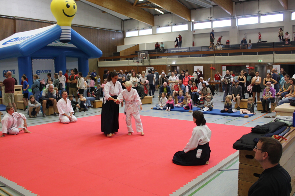 Aikidoka bei einer Vorführung. Im Hintergrund zuschauende Menschen und eine Hüpburg.