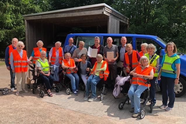 Jens Deichm&ouml;ller und Rainer Pommeranz pr&auml;stentieren vor dem Bus umringt von den Teilnehmern der Rollatorgruppe stolz ihr Urkunden zur Auszeichnung zum moin!Vereinshelden