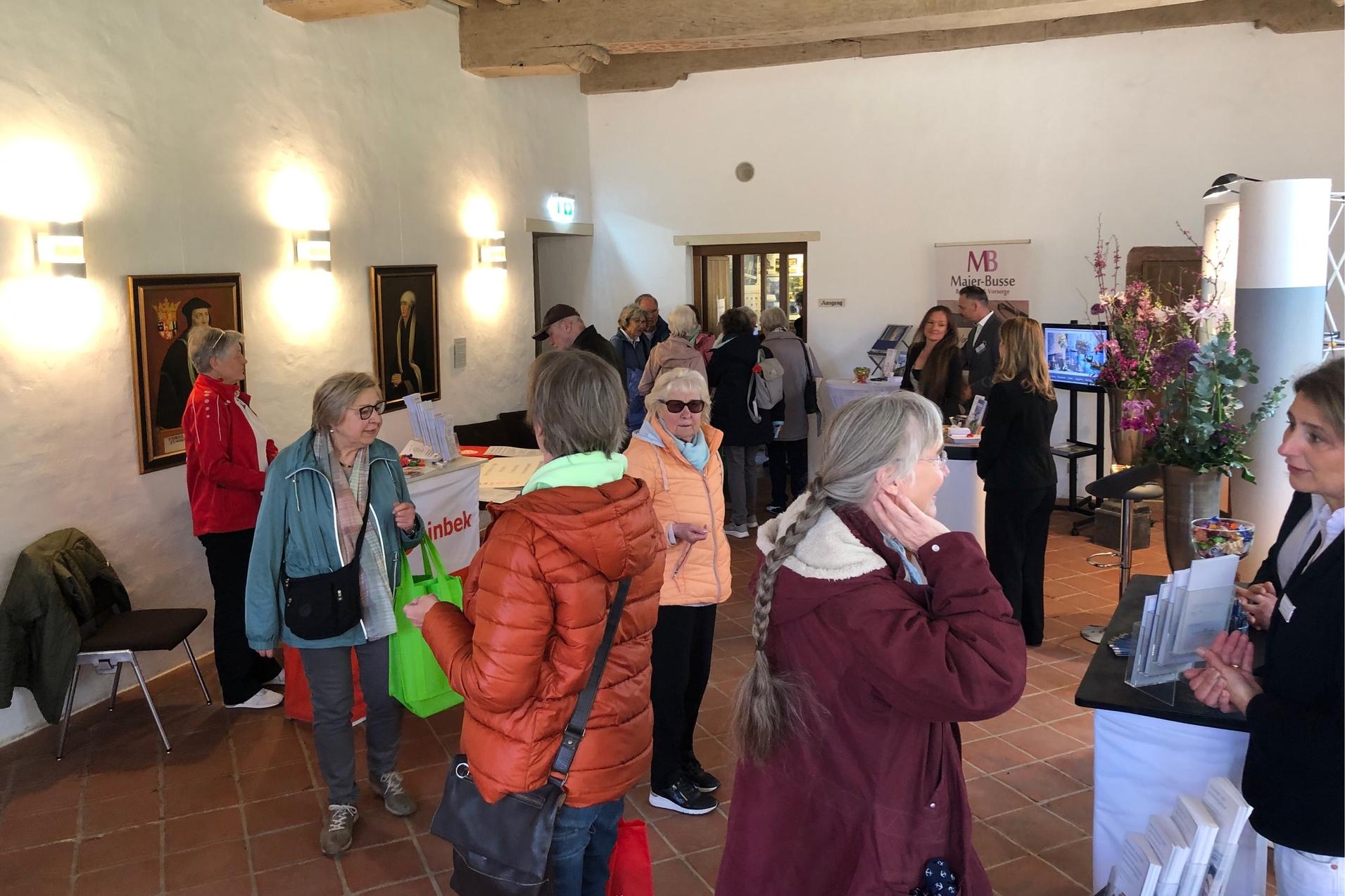 Viele Personen stehen in einem Raum an verschiedenen St&auml;nden und unterhalten sich