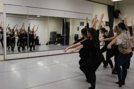 Gruppe Tänzerinnen in schwarzer Sportkleidung tanzt mit ausgebreiteten Armen vor einem Spiegel.