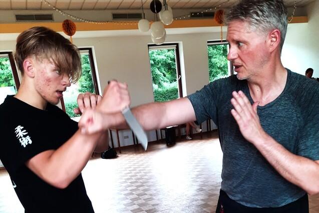 2 Erwachsene Personen simulieren Nahkampfsituation. Einer ist mit einem Messer bewaffnet. Der anderer schnellt mit seiner Faust zum Arm seines Gegners