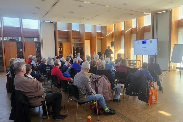 Publikum sitzt auf den St&uuml;hlen und h&ouml;rt den Worten des Referenten zu