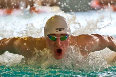 Schwimmer mit weißer Badekappe und weit geöffnetem Mund. Die Arme tauchen hinter den Schultern aus dem Wasser auf.