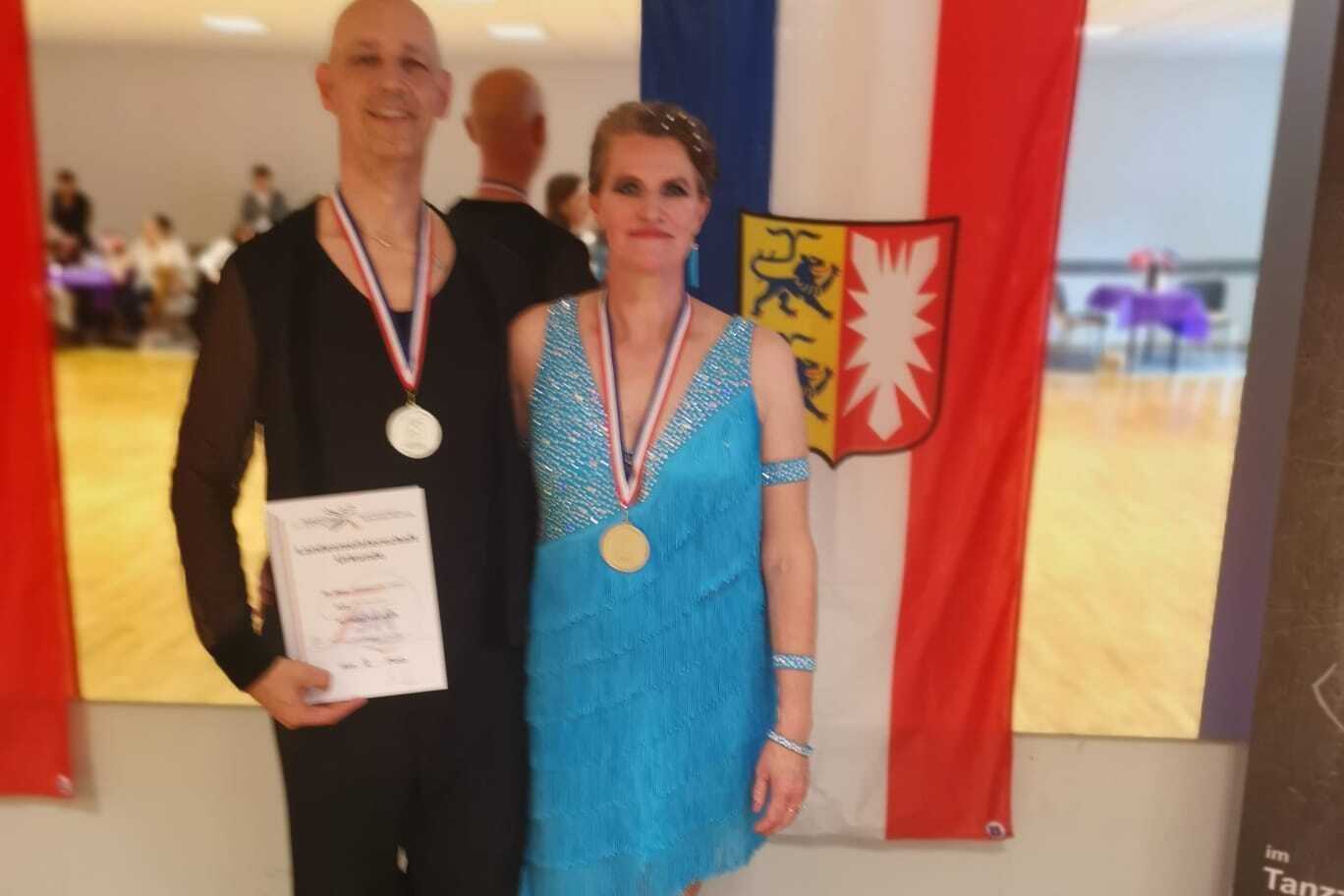 Ehepaar Schuldt ertanzt Silbermedaillen Ehepaar Schuldt präsentiert Arm in Arm vor einer Spiegelwand mit schleswig-holsteinischer Landesfahne ihre Urkunde und Silbermedaille.
