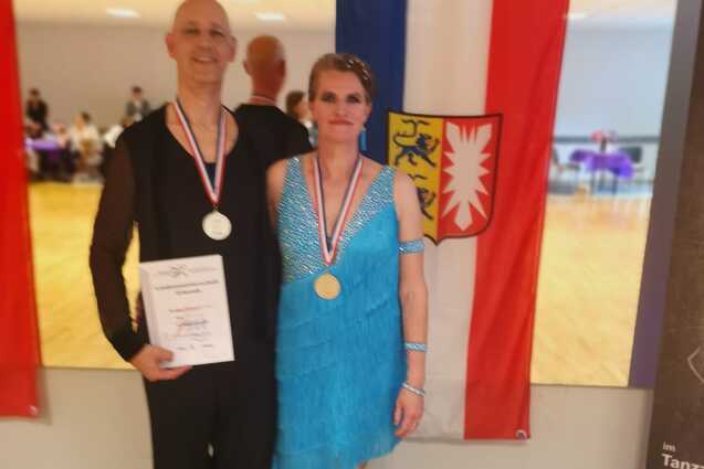 Ehepaar Schuldt pr&auml;sentiert Arm in Arm vor einer Spiegelwand mit schleswig-holsteinischer Landesfahne ihre Urkunde und Silbermedaille.