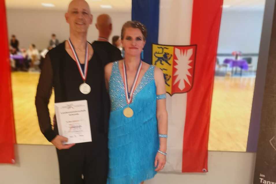 Ehepaar Schuldt ertanzt Silbermedaillen Ehepaar Schuldt präsentiert Arm in Arm vor einer Spiegelwand mit schleswig-holsteinischer Landesfahne ihre Urkunde und Silbermedaille.