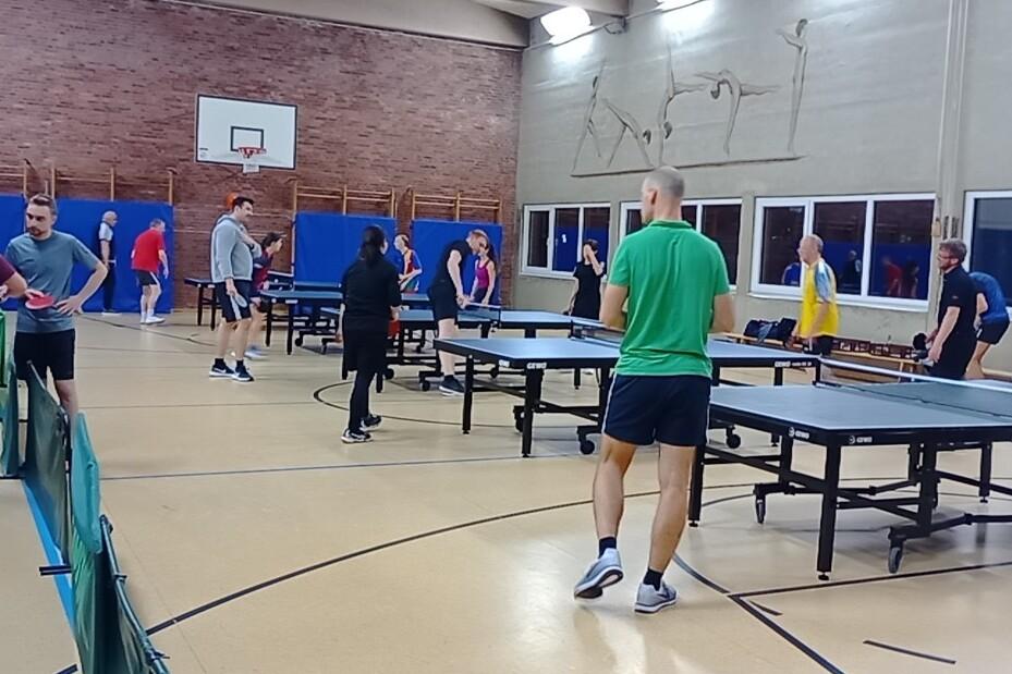 TischtennisspielerInnen an Platten. Daneben stehen Spieler und unterhalten sich.