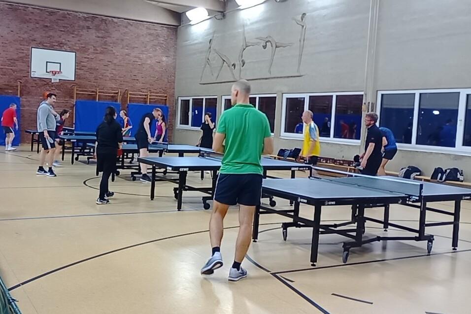 TischtennisspielerInnen an Platten. Daneben stehen Spieler und unterhalten sich.