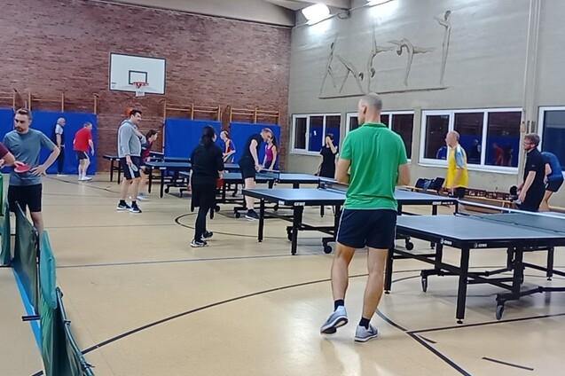 TischtennisspielerInnen an Platten. Daneben stehen Spieler und unterhalten sich.