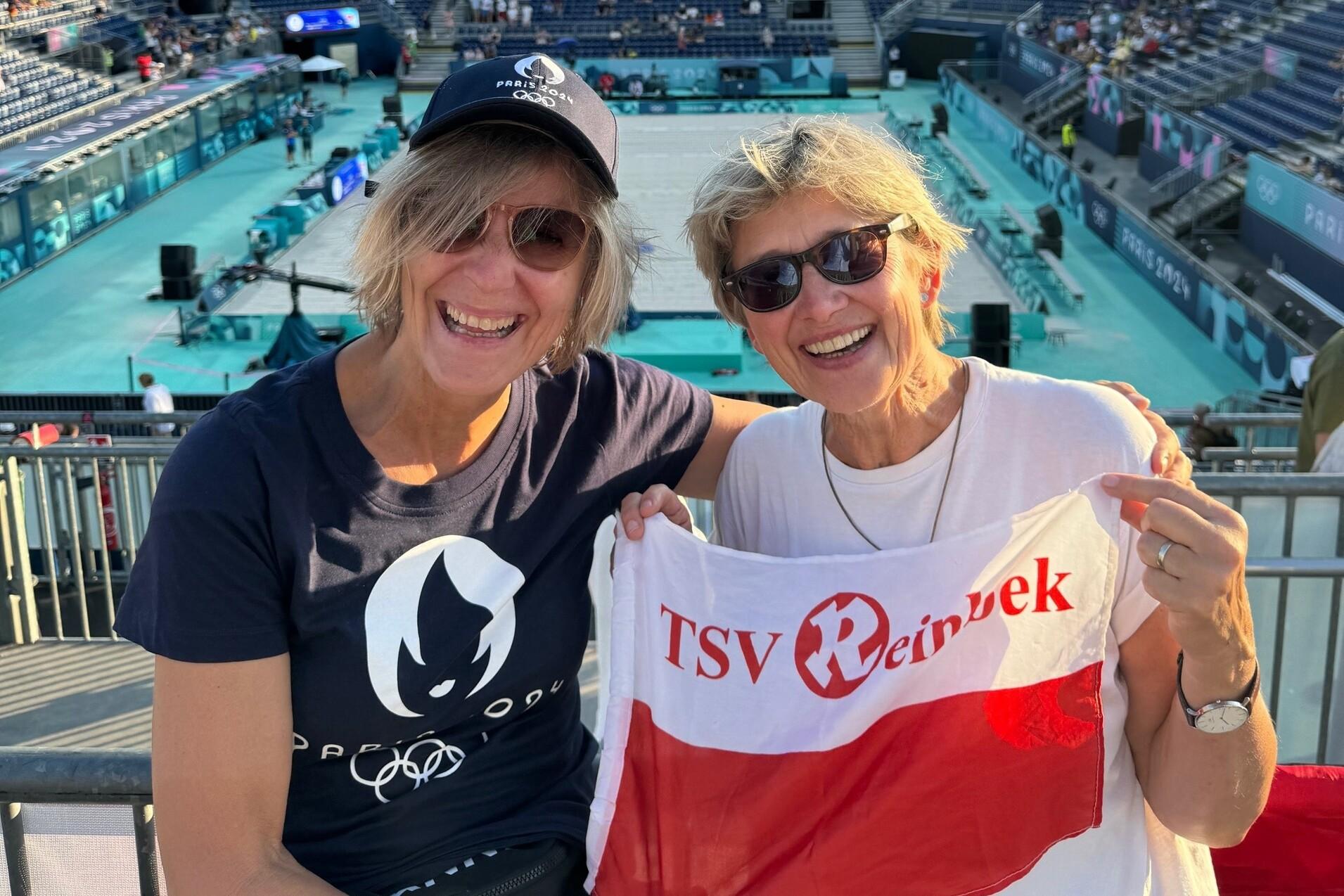 Anni und Maren bei den olympischen Spielen 2024 Volleyball Freizeitsportlerinnen Anni und Maren vor dem Volleyballfeld der olympischen Spiele halten TSV Reinbek Flagge in die Kamera