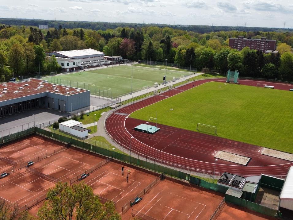 Luftaufnahme eines Sportgeländes mit Tennisplätzen, Rasensportfeld und Laufbahn, umgeben von Bäumen.