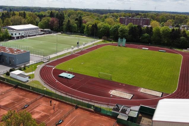 Luftaufnahme eines Sportgel&auml;ndes mit Tennispl&auml;tzen, Rasensportfeld und Laufbahn, umgeben von B&auml;umen.