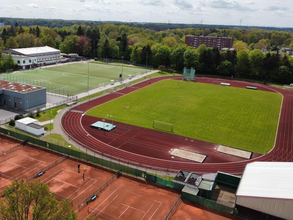 Luftaufnahme eines Sportgeländes mit Tennisplätzen, Rasensportfeld und Laufbahn, umgeben von Bäumen.