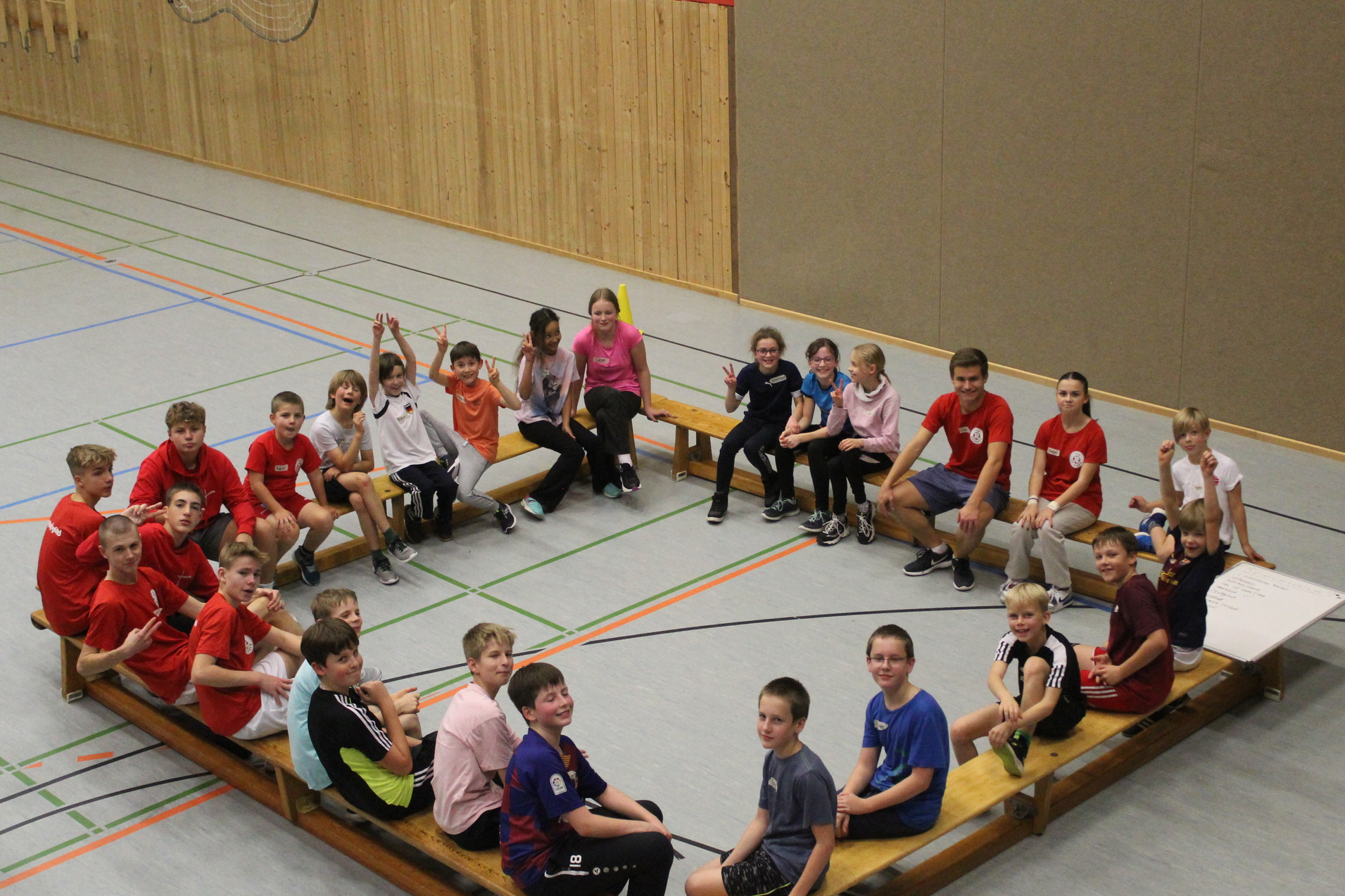 Ungefähr 20 Kinder sitzen auf Bänken die in einem Viereck stehen.