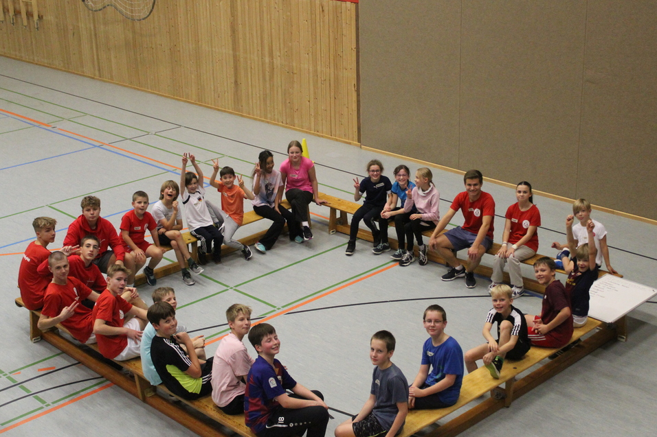 Ungefähr 20 Kinder sitzen auf Bänken die in einem Viereck stehen.