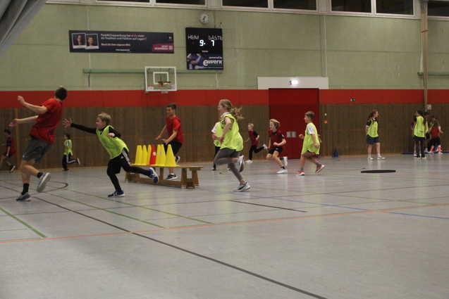 Drei Spieler der roten Mannschaft versuchen, der gelben Mannschaft ein Hütchen von ihrer Turnbank zu klauen. Die gelbe Mannschaft versucht, die roten Spieler zu ticken.