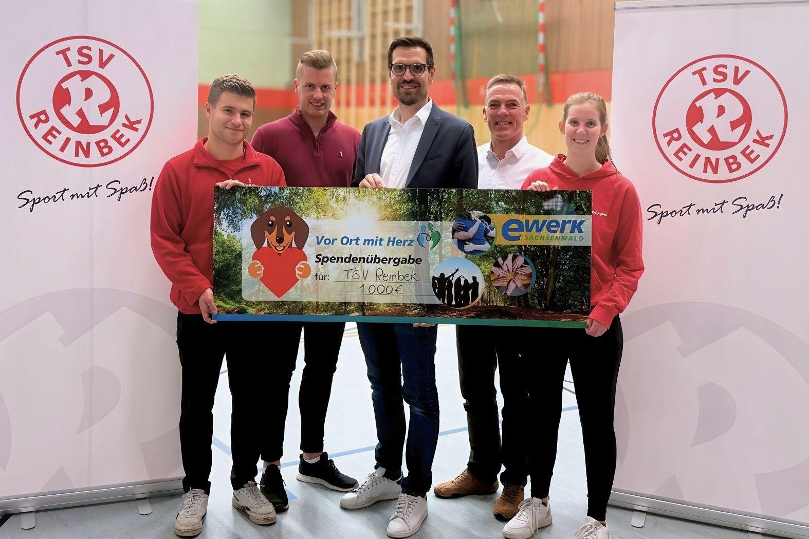 5 Personen, die sich f&uuml;r ein Gruppenfoto nebeneinander positioniert haben. Sie halten einen symbolischen Scheck in H&ouml;he von 1.000 &euro; in der Hand. Rechts und links stehen wei&szlig;e Rollups mit dem Logo der TSV Reinbek.