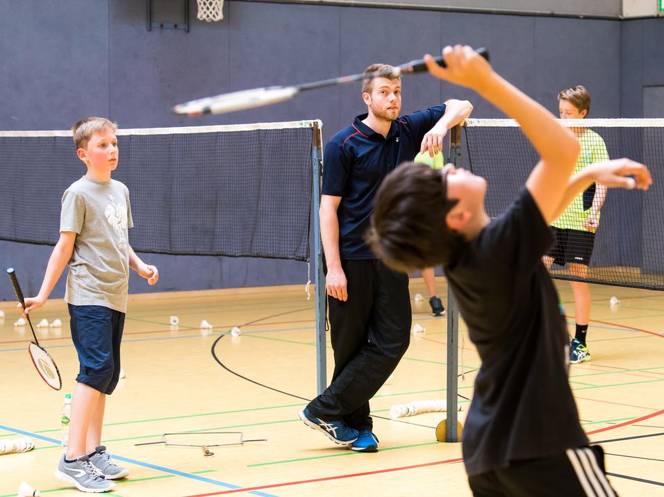 Gruppentraining im Badminton: Kinder üben mit Schlägern und Bällen unter Anleitung eines Trainers in einer Sporthalle.