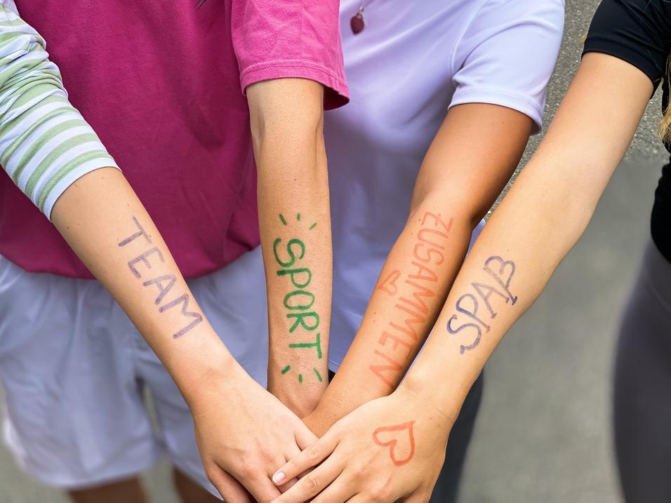 Vier Arme liegen übereinander, mit farbiger Schrift: „TEAM“, „SPORT“, „ZUSAMMEN“, „SPASS“ auf der Haut.