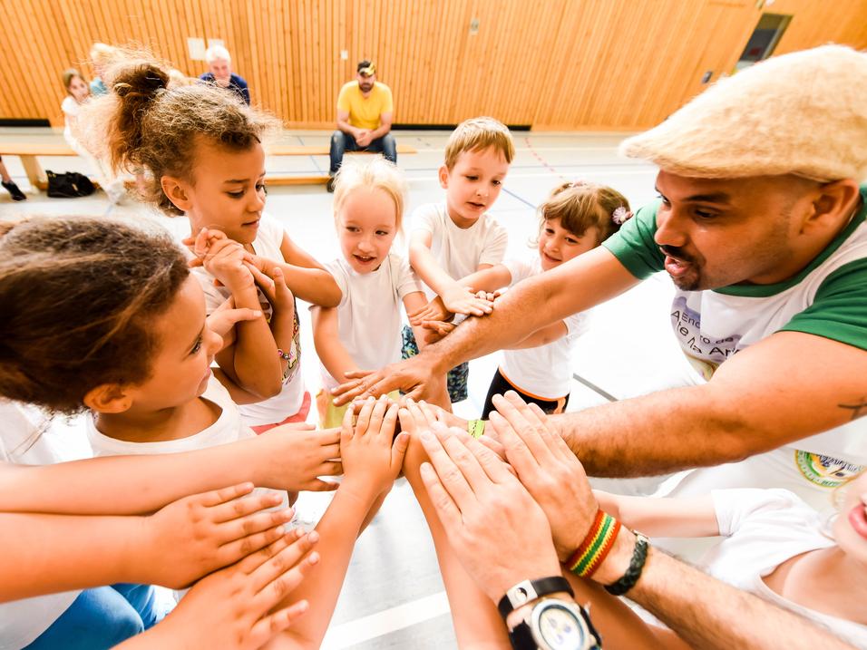 Gruppenspiel mit Kindern, die ihre Hände zum gemeinsamen Aufeinandertreffen heben, unter Anleitung eines Erwachsenen.