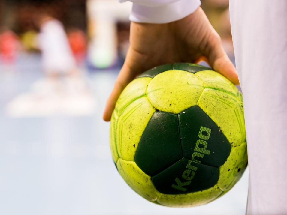 Nahaufnahme einer Hand, die einen grünen Handball mit gelben Elementen hält, im Hintergrund Spieler auf dem Spielfeld.