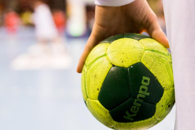 Nahaufnahme einer Hand, die einen grünen Handball mit gelben Elementen hält, im Hintergrund Spieler auf dem Spielfeld.