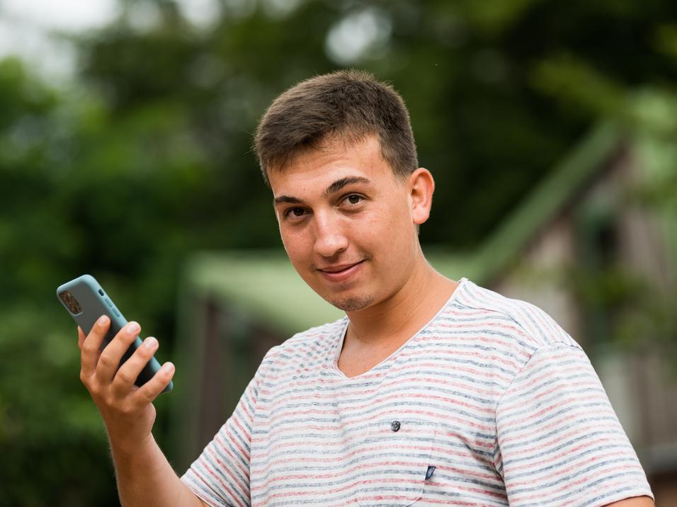 Junger Mann mit kurzen Haaren hält ein Smartphone in der Hand und schaut lächelnd in die Kamera, Hintergrund unscharf.