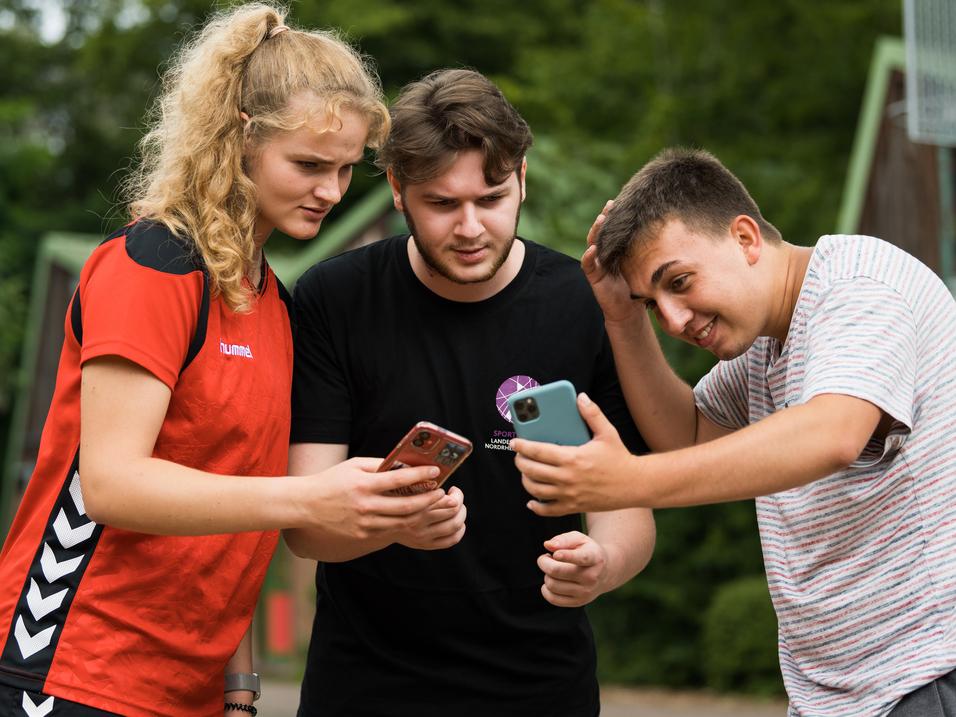 Drei Jugendliche stehen zusammen und schauen auf ihre Smartphones, um etwas anzusehen oder zu besprechen.