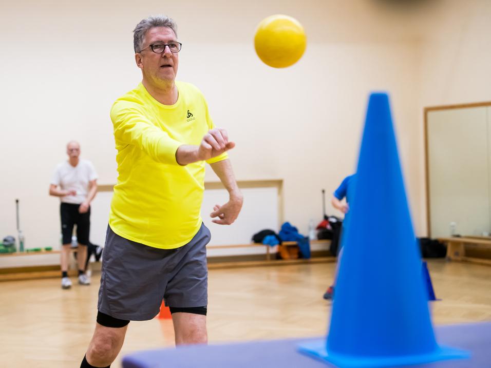 Ein Mann in gelber Sportkleidung wirft einen Ball auf einen blauen Kegel in einem Übungsraum. Hintergrund unscharf.