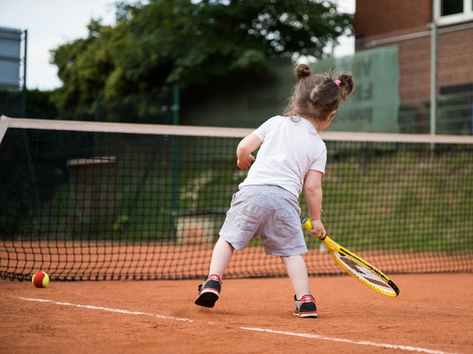 Kind mit Zöpfen schlägt Tennisball auf einem Sandplatz, während es sich auf einen Schlag vorbereitet.