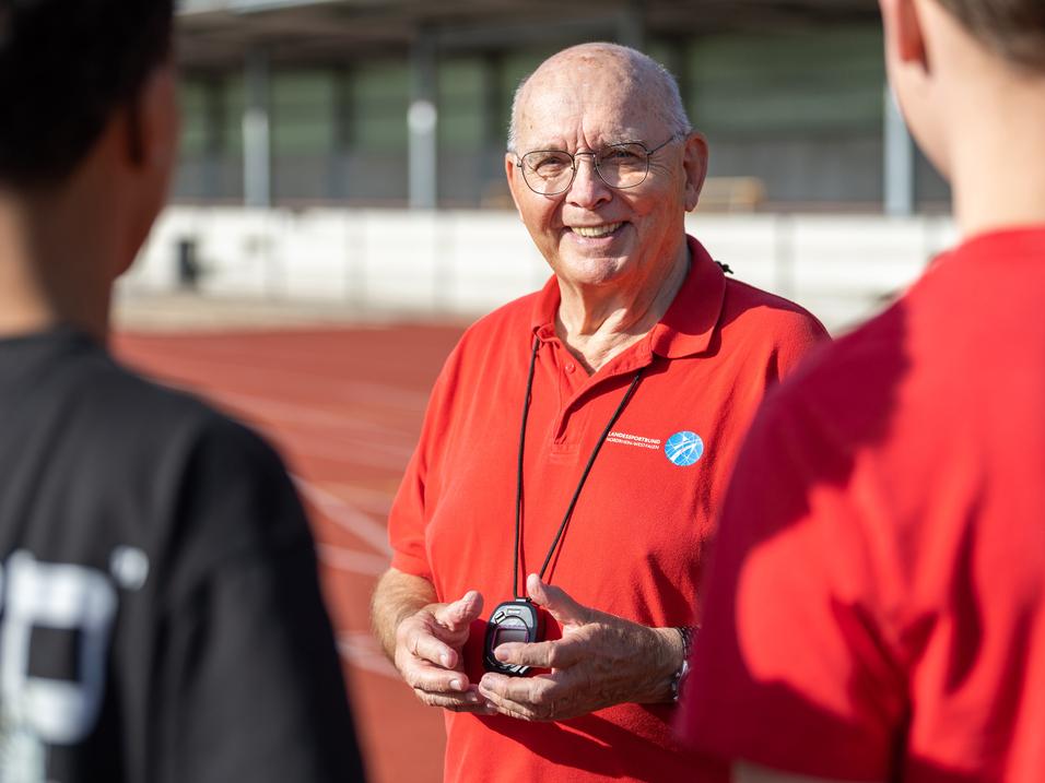 Ein älterer Mann in rotem Shirt mit Stoppuhr spricht an einer Laufbahn mit zwei Jugendlichen.