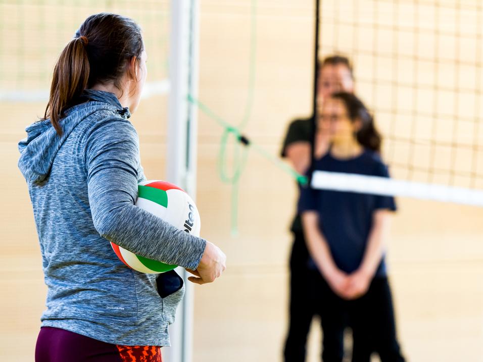 Eine Person mit einem Volleyball steht vor dem Netz, während zwei weitere Personen im Hintergrund warten.